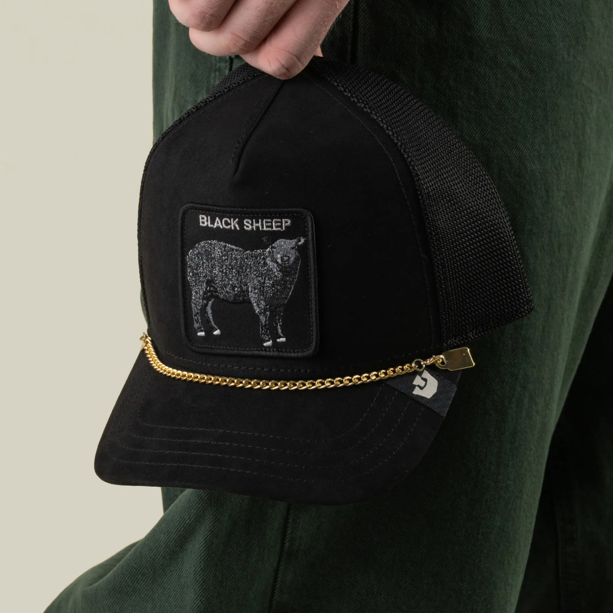 A hand holds a black mesh trucker hat by Goorin Bros., featuring a BLACK SHEEP patch, a sheep graphic, and the stylish Mini CapLoc Chain—hat jewelry with a gold chain and double clip chains adorning the front. The person is wearing dark green pants.