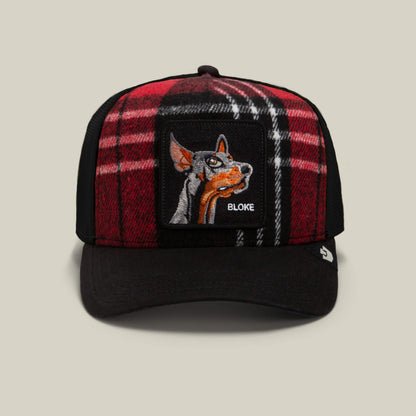 A red-and-black plaid trucker hat from Goorin Bros., the “Brit Bloke,” featuring an embroidered patch with a detailed dog’s head on the front.