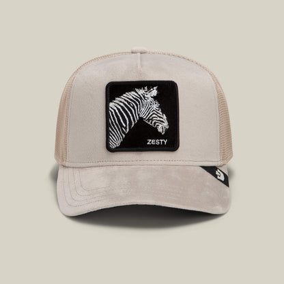 The Velour Zesty trucker hat by Goorin Bros. features a beige color with a mesh back, and showcases a front patch of a zebra head with monochrome stripes and the word “ZESTY.”.