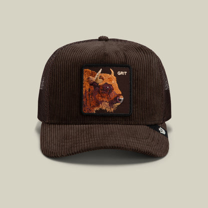 The Goorin Bros. Corduroy Grit trucker hat features dark brown corduroy fabric and a breathable mesh back, accented by a stitched patch showcasing a bison and the word “GRIT” on the front. It is finished with a curved brim for classic style.