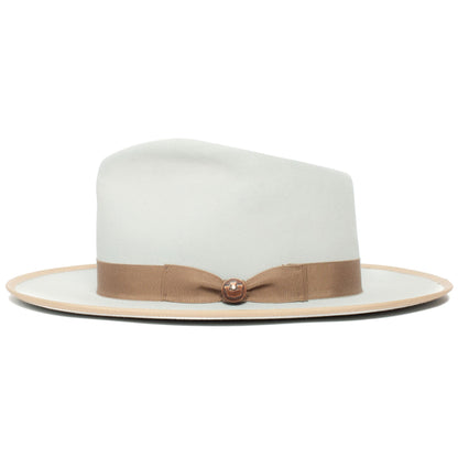 The River Gray by Goorin Bros. is a light gray merino wool fedora with a wide brim, accented by a beige ribbon band and a small round decorative pin on the band, displayed on a white background. Made in America, this wide brim fedora offers timeless style and subtle elegance.
