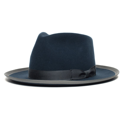 River Gray by Goorin Bros. is a dark blue merino wool fedora featuring a black ribbon band and a flat, slightly upturned brim, displayed against a white background. This American-made wide brim fedora exudes timeless sophistication.