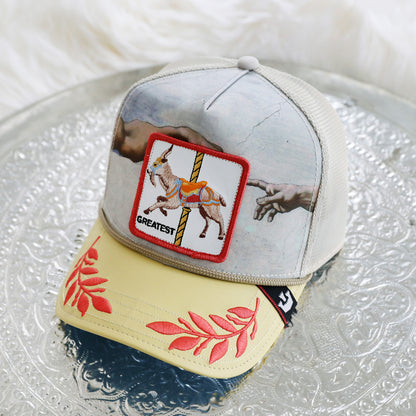 A The Farm “Maximum” mesh cap, featuring a printed mural of hands, a “Greatest” goat patch, and red leaf embroidery on the yellow brim, rests on a patterned silver tray. *Please note: All Sales Are Final.*.